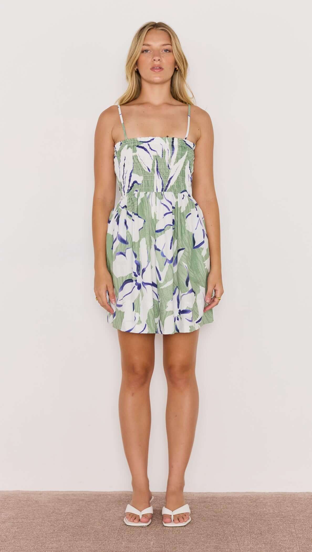 Model wearing a green and white shirred mini dress with floral design and sandals, displayed against a plain background.