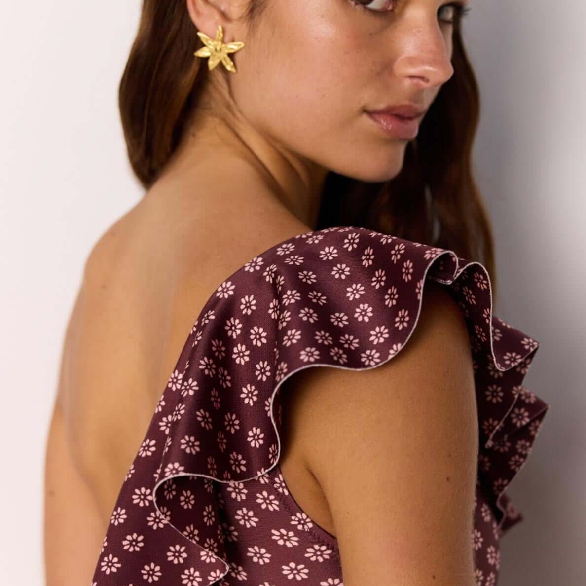 Model wearing a burgundy floral print one-shoulder swimsuit with ruffle detail, showcasing summer fashion.