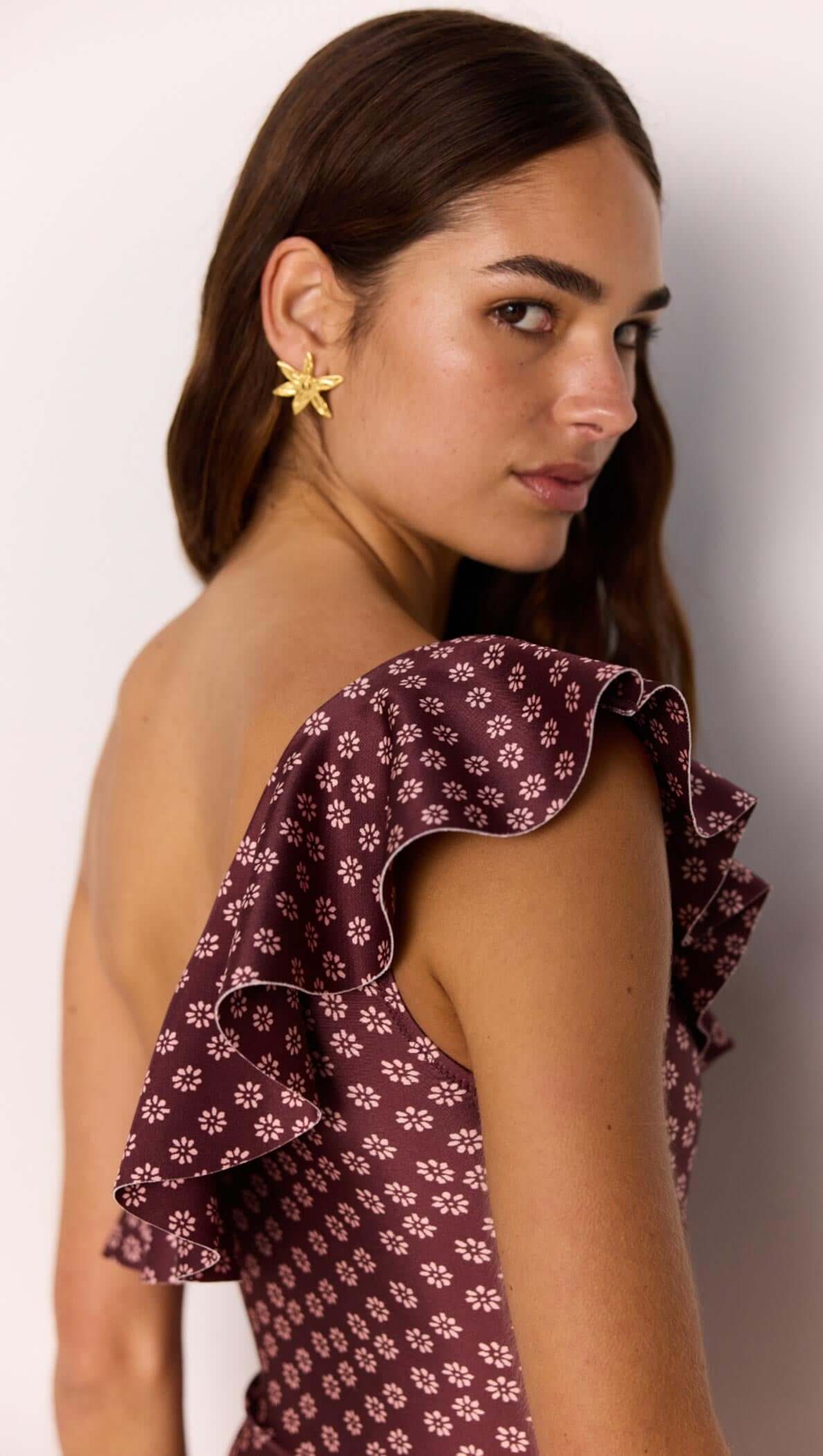 Model wearing a burgundy floral print one-shoulder swimsuit with ruffle detail, showcasing summer fashion.