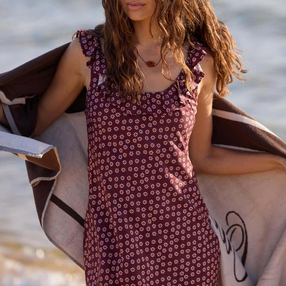 Model wearing a Burgundy Floral Print Ruffle Strap Low-Back Mini Dress at the beach, holding a towel.