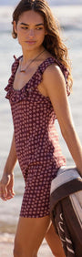 Young woman wearing a ruffle strap burgundy floral mini dress, walking on the beach with a towel.