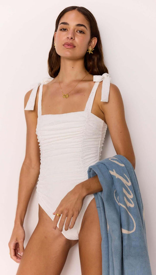 Model in a square-neck white ruched one-piece swimsuit with tie straps, holding a blue towel.