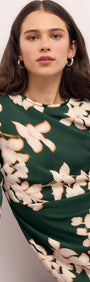 Woman modeling the Green & Cream Floral Satin Mini Dress with a forest green background and floral design accents.