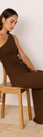 One-Shoulder Brown Midi Dress with Beaded Belt worn by model sitting on a chair, showcasing its elegant design and silhouette.